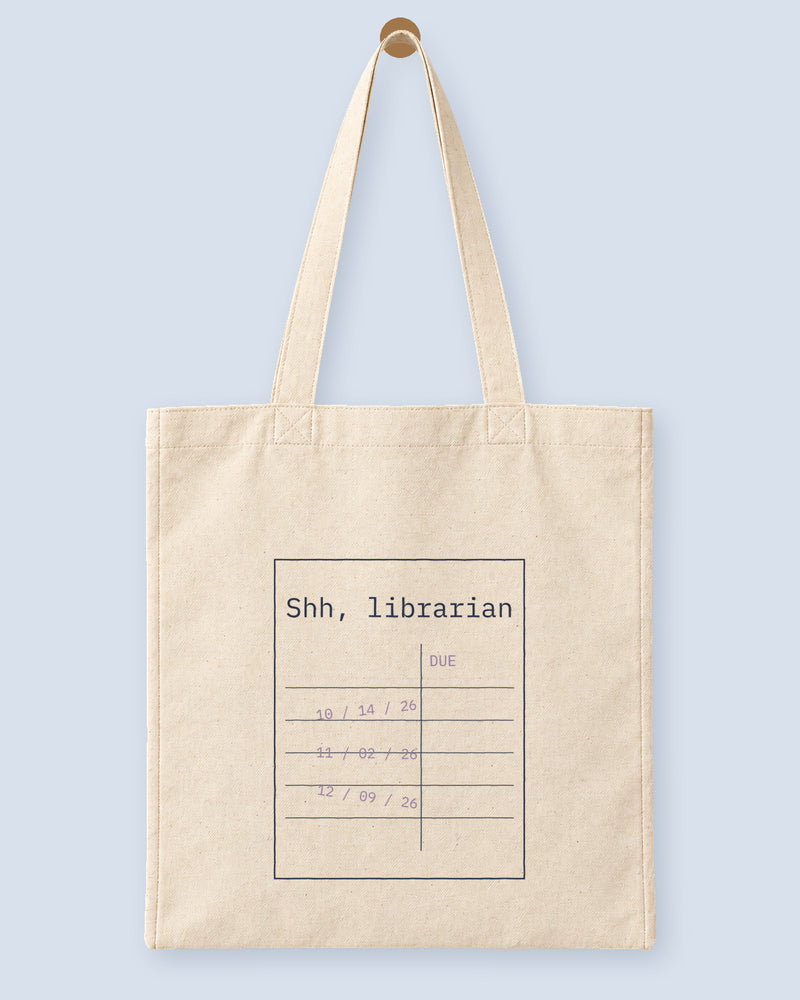 Natural canvas tote printed with “Shh, librarian”, hanging on a wooden peg.