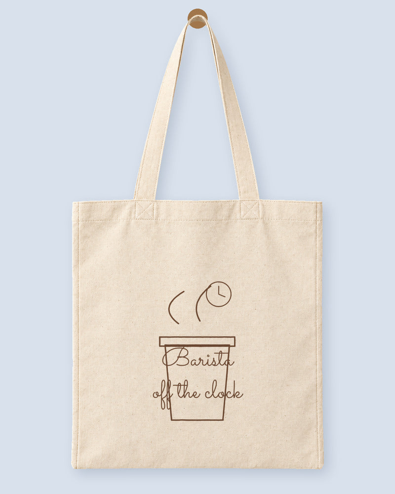 Natural canvas tote printed with “Barista off the clock”, hanging on a wooden peg.