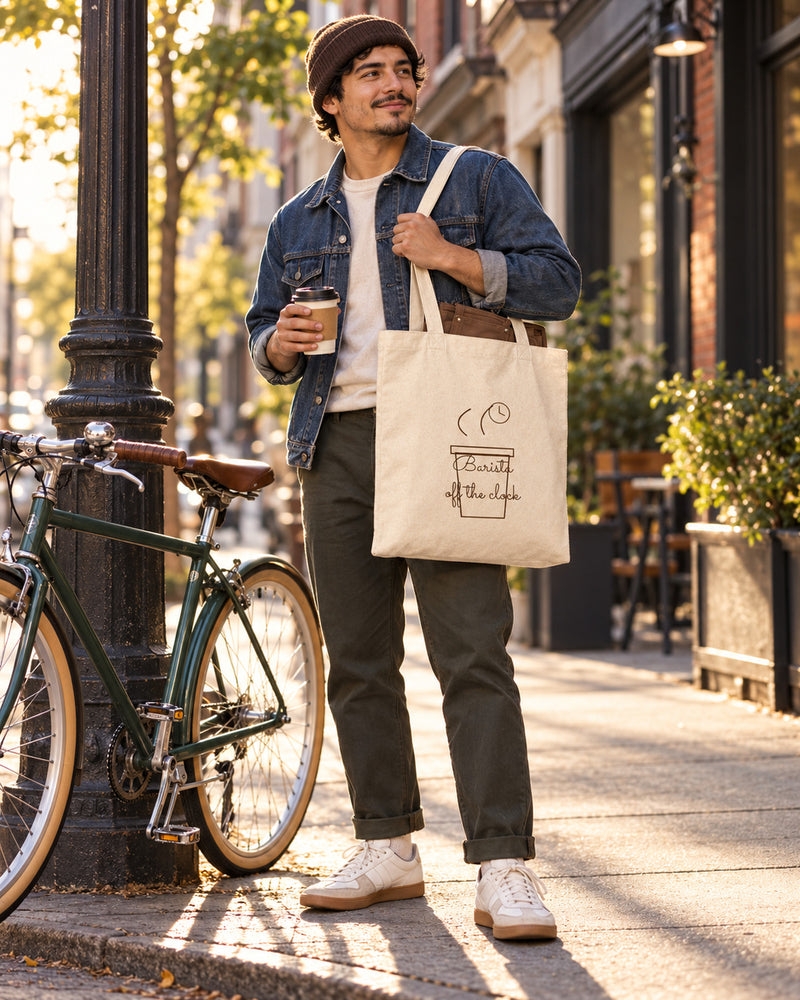Latino man carrying a natural canvas tote printed with “Barista off the clock” in cocoa. Styled digital image.