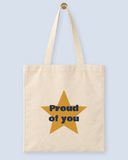 Natural canvas tote printed with “Proud of you”, hanging on a wooden peg.
