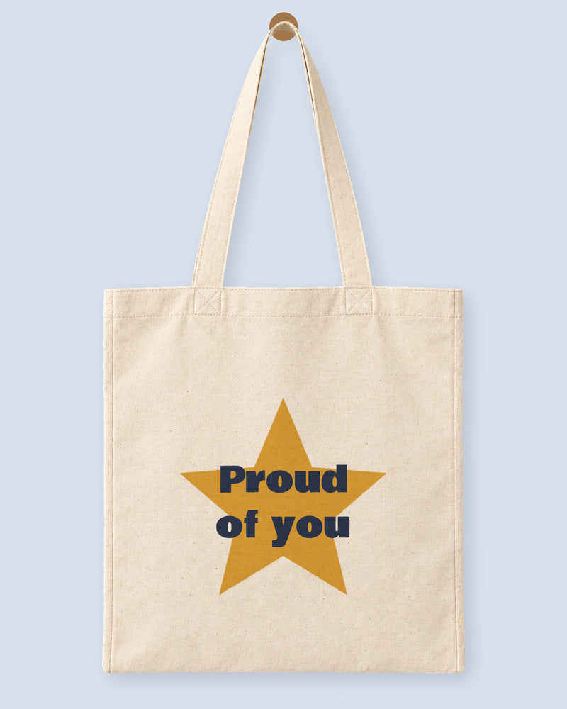 Natural canvas tote printed with “Proud of you”, hanging on a wooden peg.