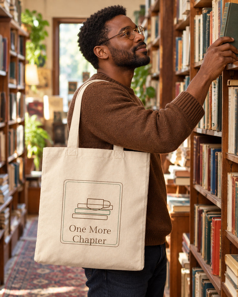 Black man carrying a natural canvas tote printed with “One More Chapter” in cocoa, forest. Styled digital image.