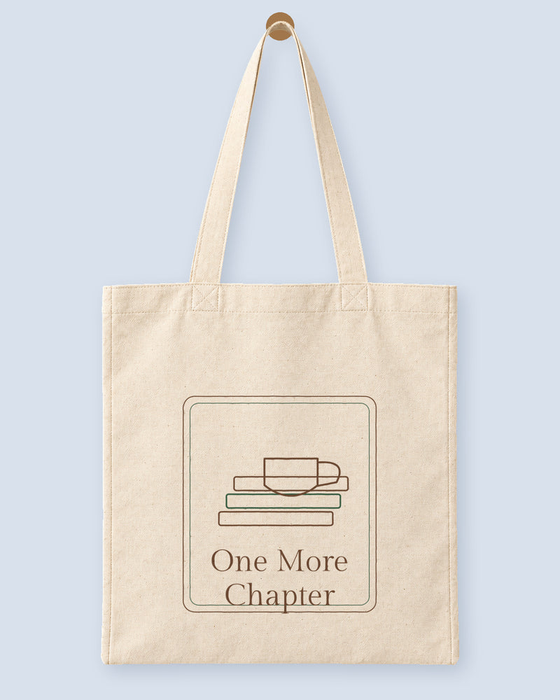 Natural canvas tote printed with “One More Chapter”, hanging on a wooden peg.