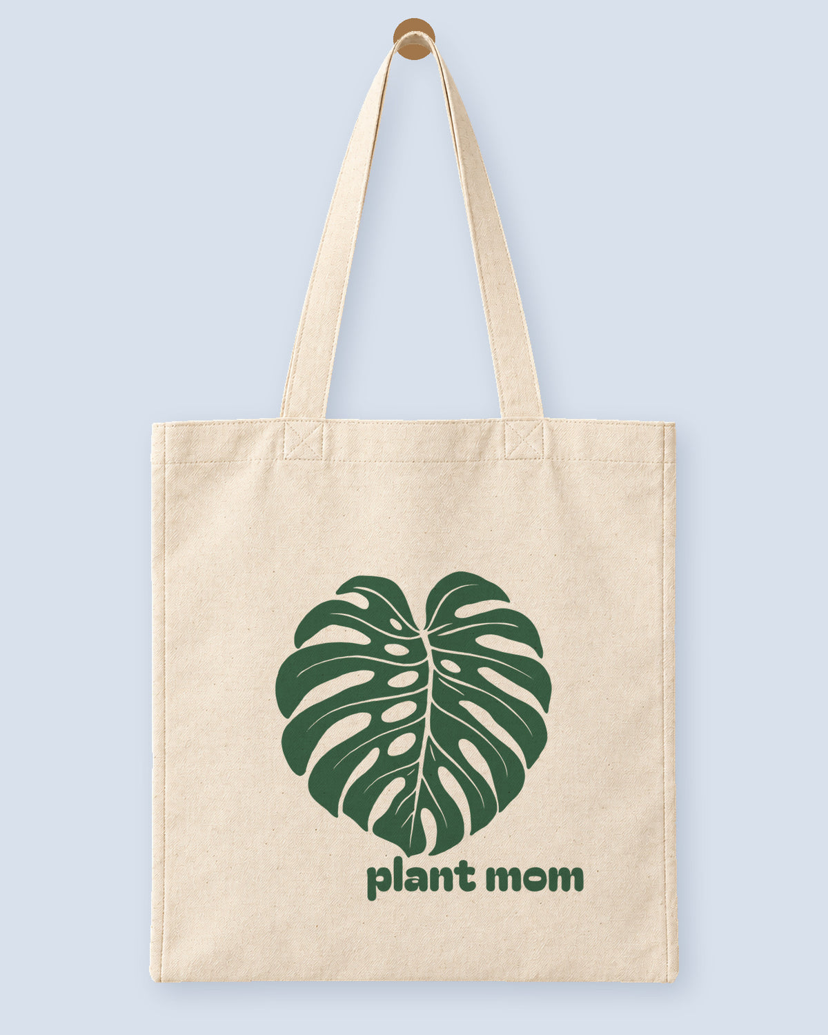 Natural canvas tote printed with “plant mom”, hanging on a wooden peg.