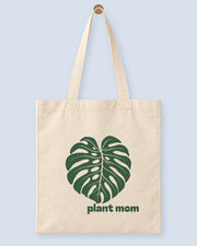 Natural canvas tote printed with “plant mom”, hanging on a wooden peg.
