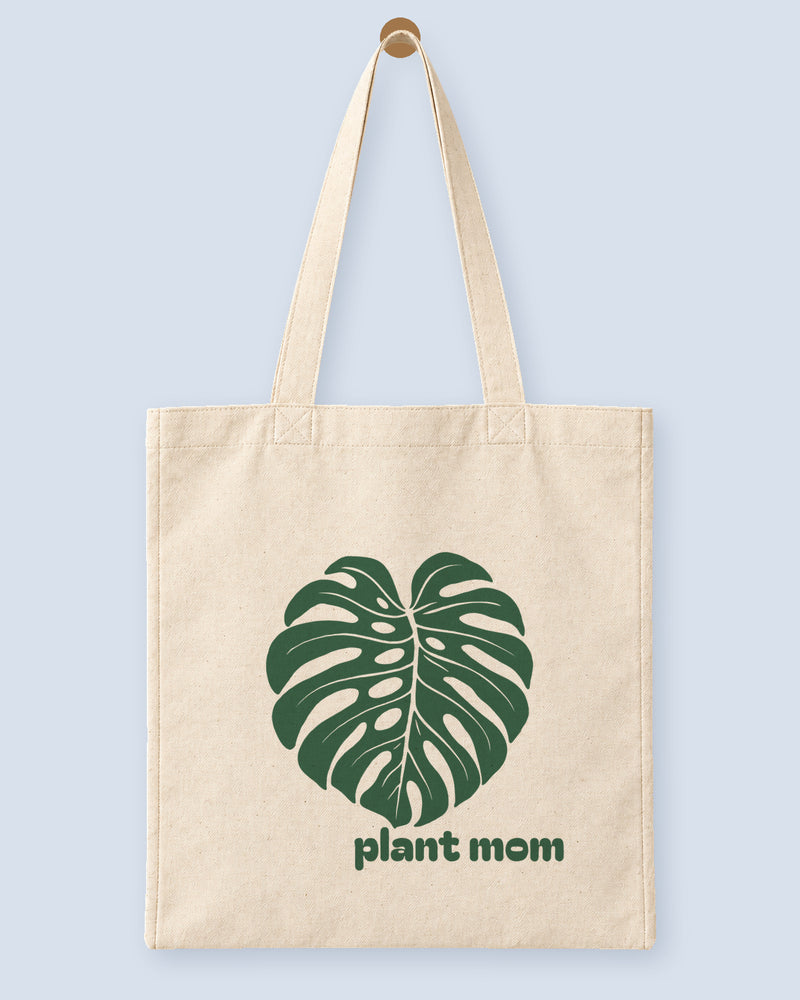 Natural canvas tote printed with “plant mom”, hanging on a wooden peg.