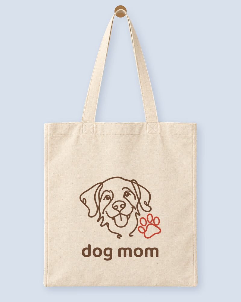 Natural canvas tote printed with “dog mom”, hanging on a wooden peg.