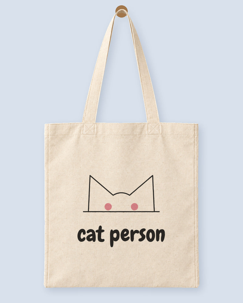 Natural canvas tote printed with “cat person”, hanging on a wooden peg.