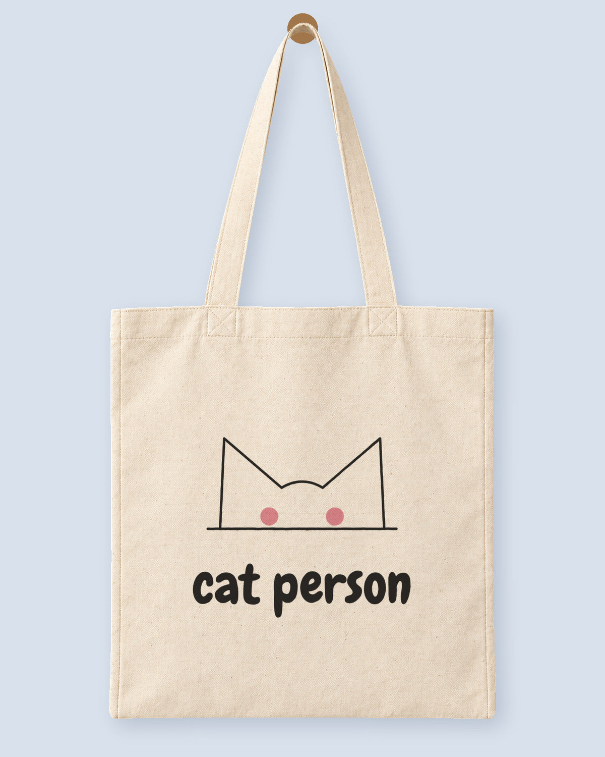 Natural canvas tote printed with “cat person”, hanging on a wooden peg.