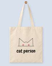Natural canvas tote printed with “cat person”, hanging on a wooden peg.