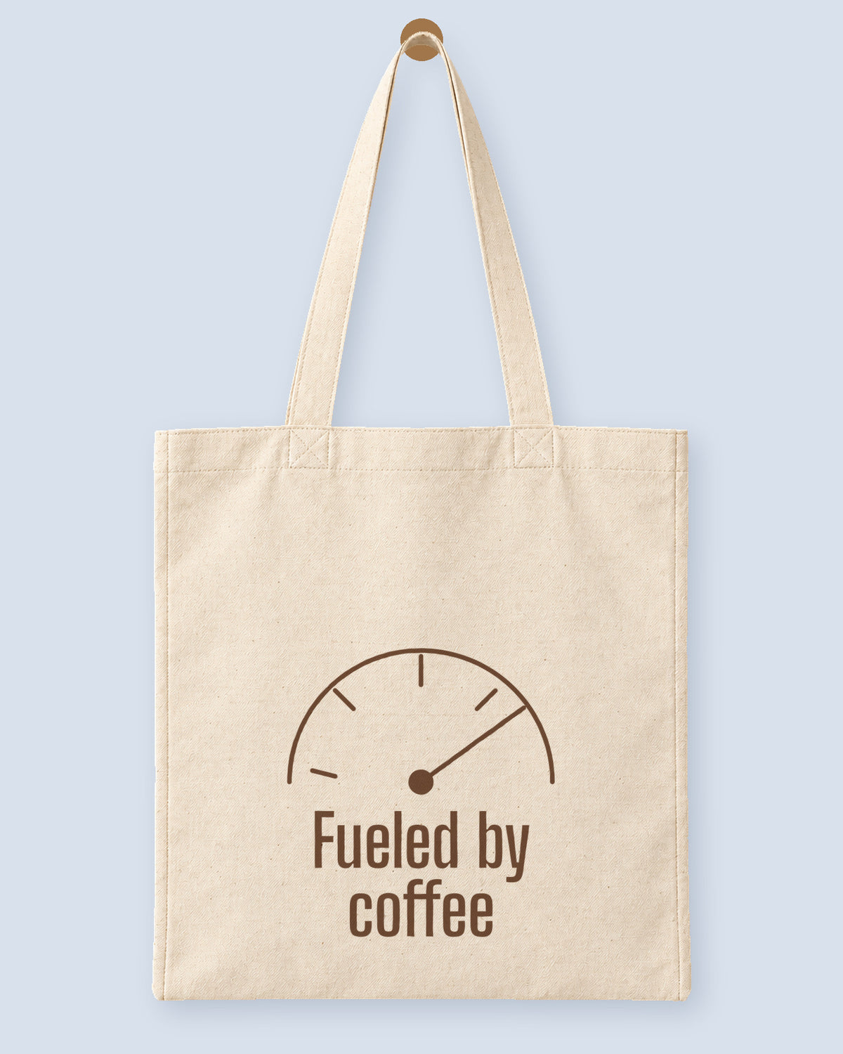 Natural canvas tote printed with “Fueled by coffee”, hanging on a wooden peg.