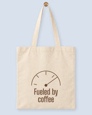 Natural canvas tote printed with “Fueled by coffee”, hanging on a wooden peg.