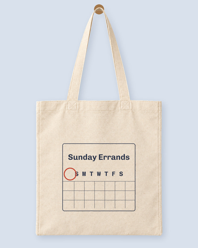 Natural canvas tote printed with “Sunday Errands”, hanging on a wooden peg.